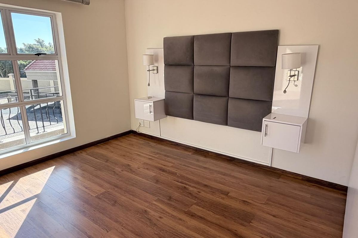 A bright, empty bedroom featuring laminate flooring, a modern upholstered headboard, and integrated wall-mounted bedside cabinetry with built-in lighting.