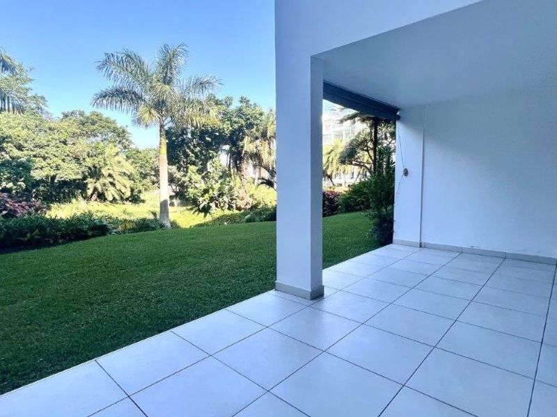 A spacious covered patio featuring clean tiled flooring, overlooking a lush green garden and mature trees.