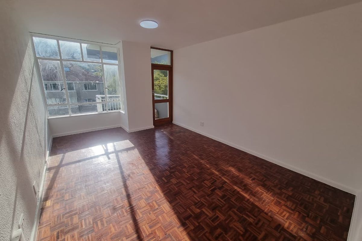 A bright, empty living room featuring classic parquet wood flooring, large windows for natural light, and direct access to a balcony.