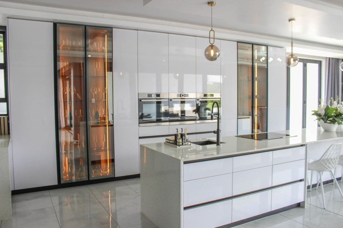 Modern kitchen featuring high-gloss white cabinetry, integrated ovens, a central island with stone countertop, and elegant glass-front display cabinets with internal lighting.