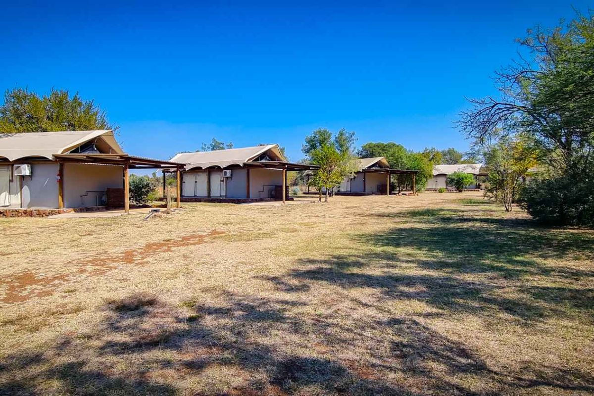 A row of detached lodge-style units featuring canvas-style roofing, timber support structures, and exterior air conditioning units set within a landscaped bushveld environment.