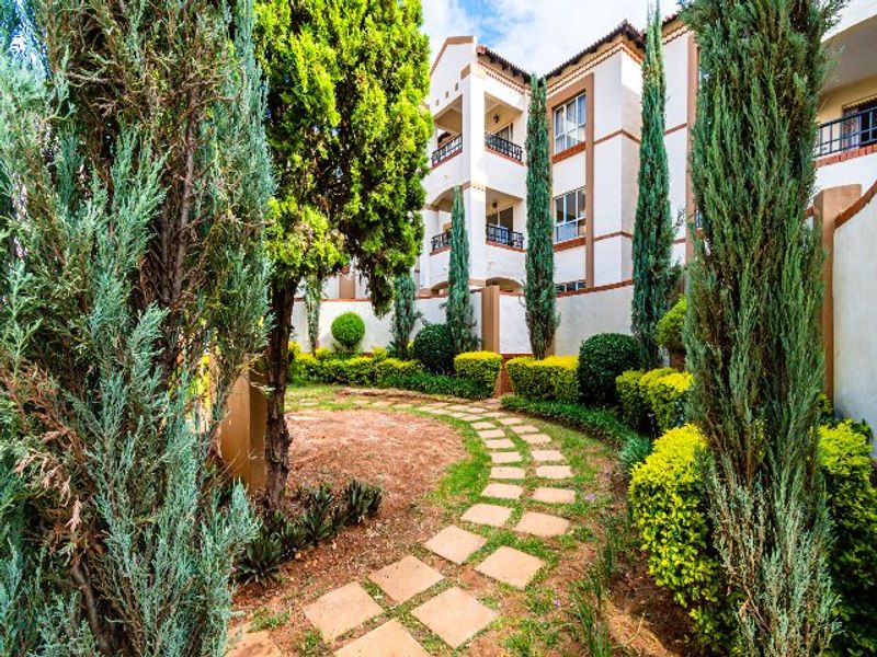 A well-maintained communal garden area featuring a stone paver pathway, manicured hedges, and tall cypress trees set against the apartment building facade.