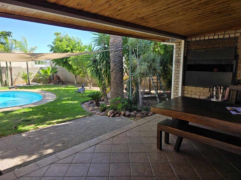 Covered patio area featuring a built-in braai, tiled flooring, and timber ceiling, overlooking a private garden with a swimming pool.