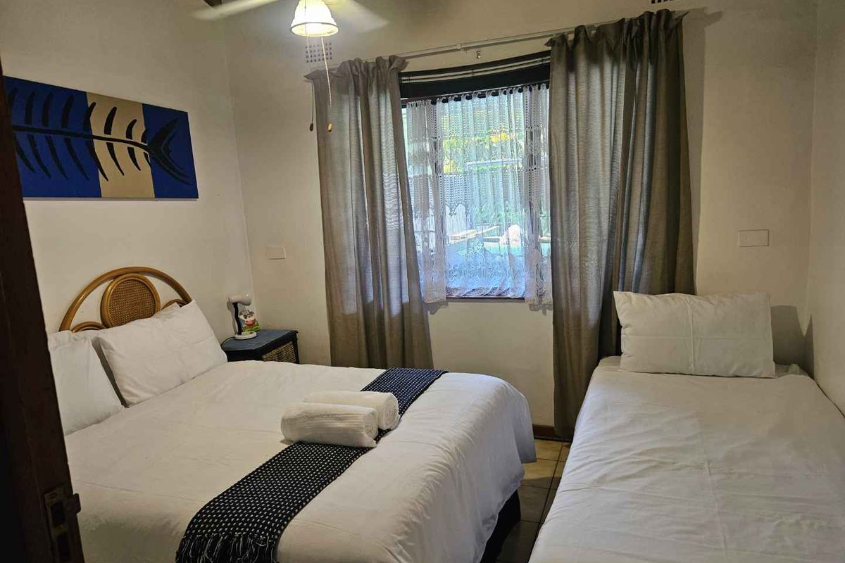 A bedroom featuring tiled flooring, a ceiling fan, and a window with curtains, providing a functional sleeping space.