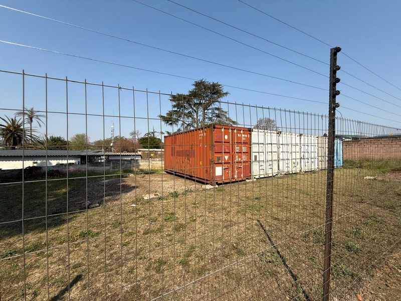 An outdoor area featuring wire mesh fencing with an electric security system, containing several shipping containers on an unpaved, undeveloped plot of land.