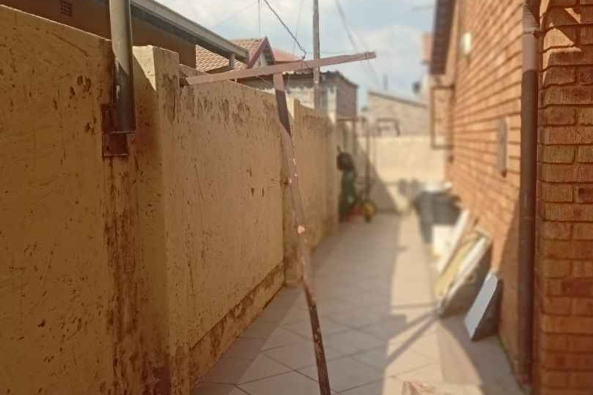 A narrow rear exterior walkway featuring tiled flooring, a brick wall, and a shade sail structure.