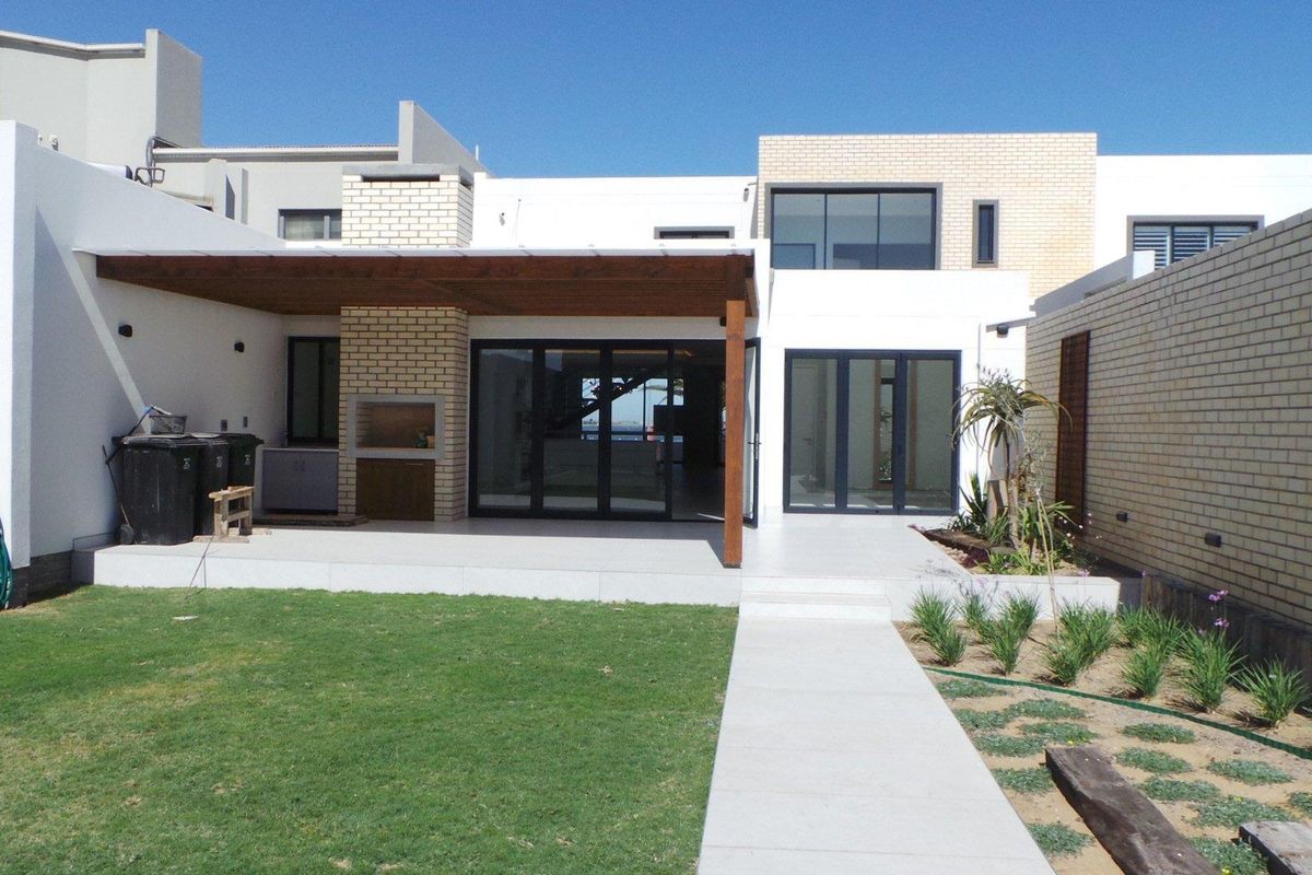 Rear view of the property featuring a covered patio with a built-in braai, tiled flooring, and a landscaped garden with a paved walkway.