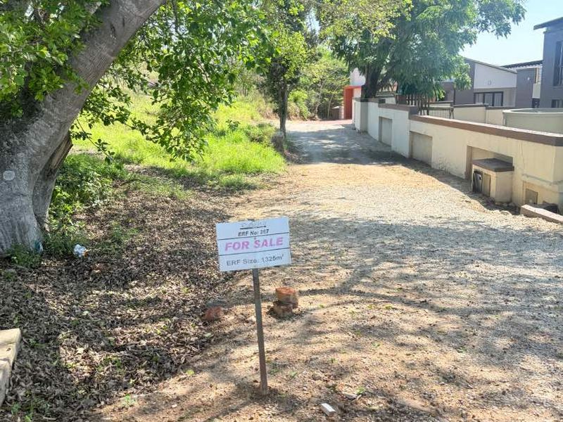 A view of a vacant residential plot featuring a gravel driveway, boundary wall, and mature trees, marked with a for sale sign.