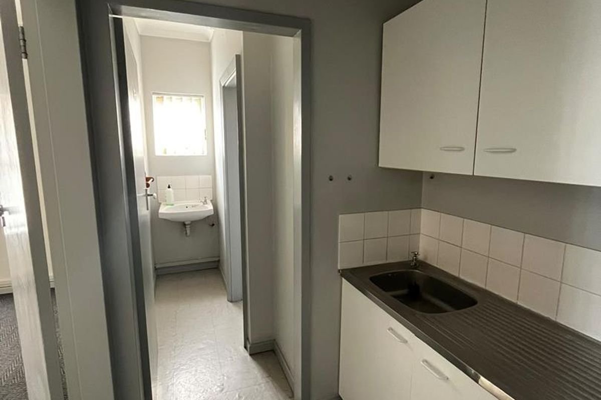 A functional kitchenette area featuring built-in cabinetry, a stainless steel sink, and tiled splashback, with an adjacent hallway leading to a washroom.