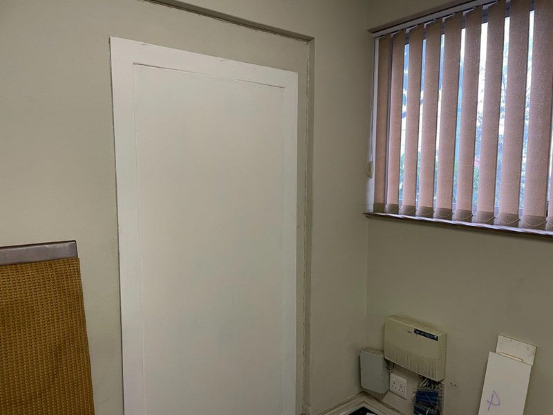 A small office space featuring vertical blinds, wall-mounted communication equipment, and exposed electrical wiring near the baseboard.