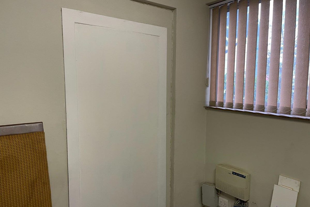A small office space featuring vertical blinds, wall-mounted communication equipment, and exposed electrical wiring near the baseboard.