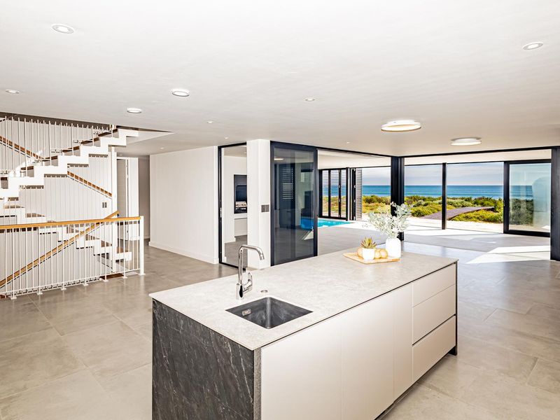 Spacious open-plan living area featuring a modern kitchen island, feature staircase, wood-burning fireplace, and seamless indoor-outdoor flow to the pool and ocean view.