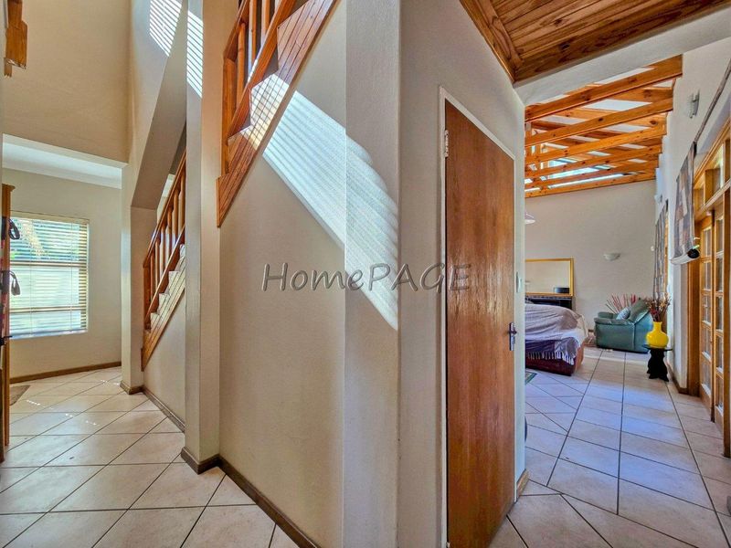 A bright hallway featuring tiled flooring, a wooden staircase, and exposed timber ceiling beams, leading into an adjacent living area.