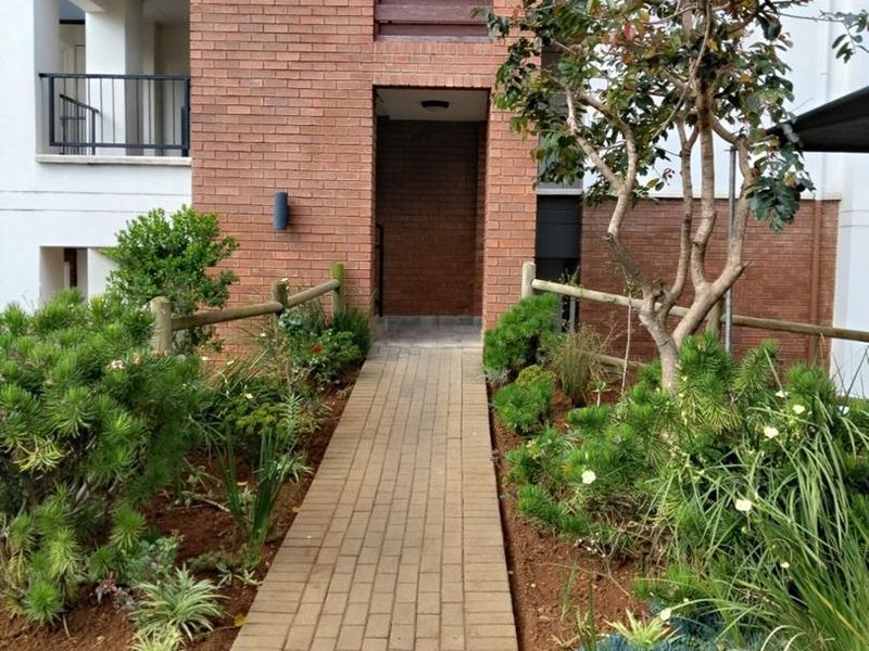 A well-maintained apartment building entrance featuring a brick facade, paved walkway, and landscaped garden area with timber fencing.
