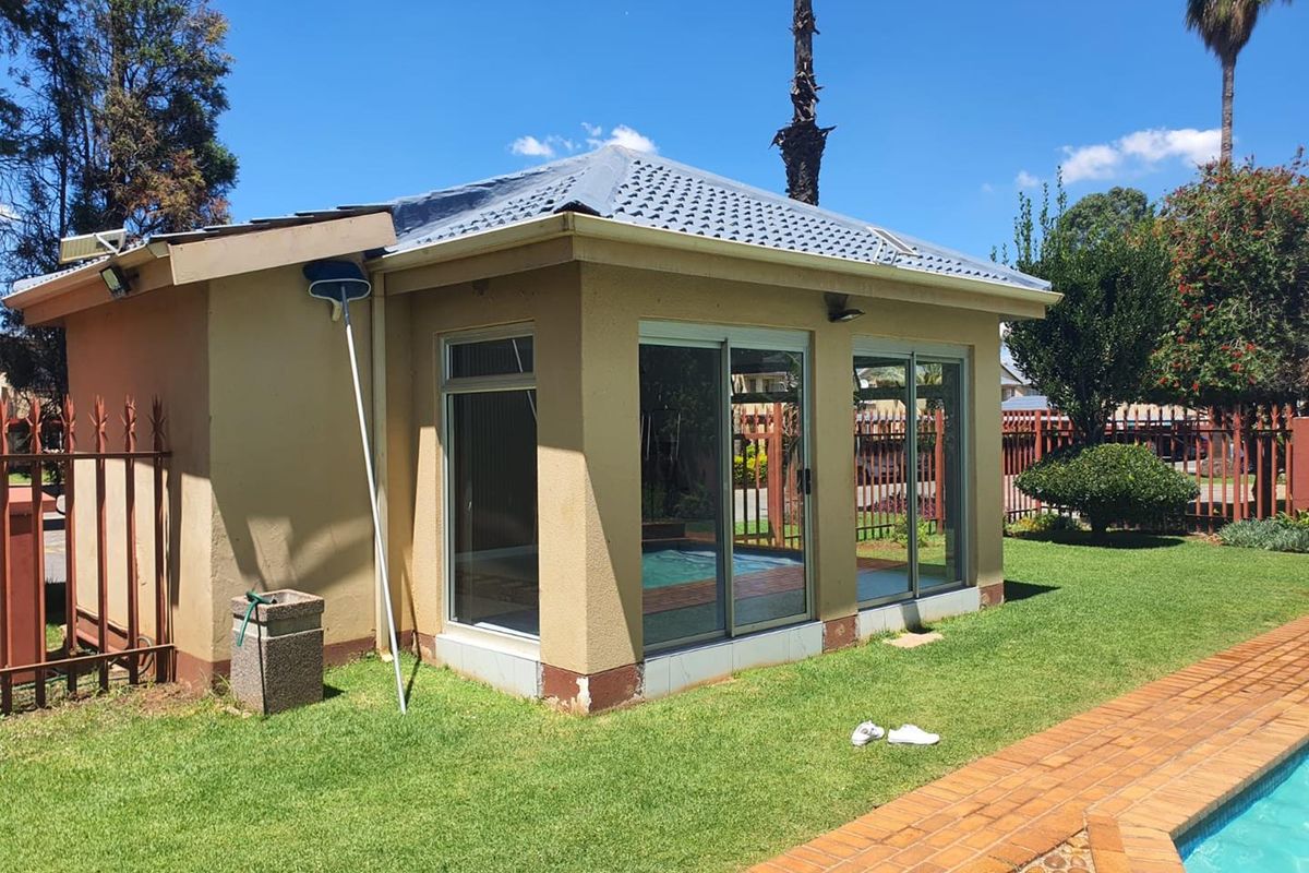 A detached garden structure featuring sliding glass doors, tiled roof, and exterior lighting, situated adjacent to a swimming pool and landscaped lawn area.