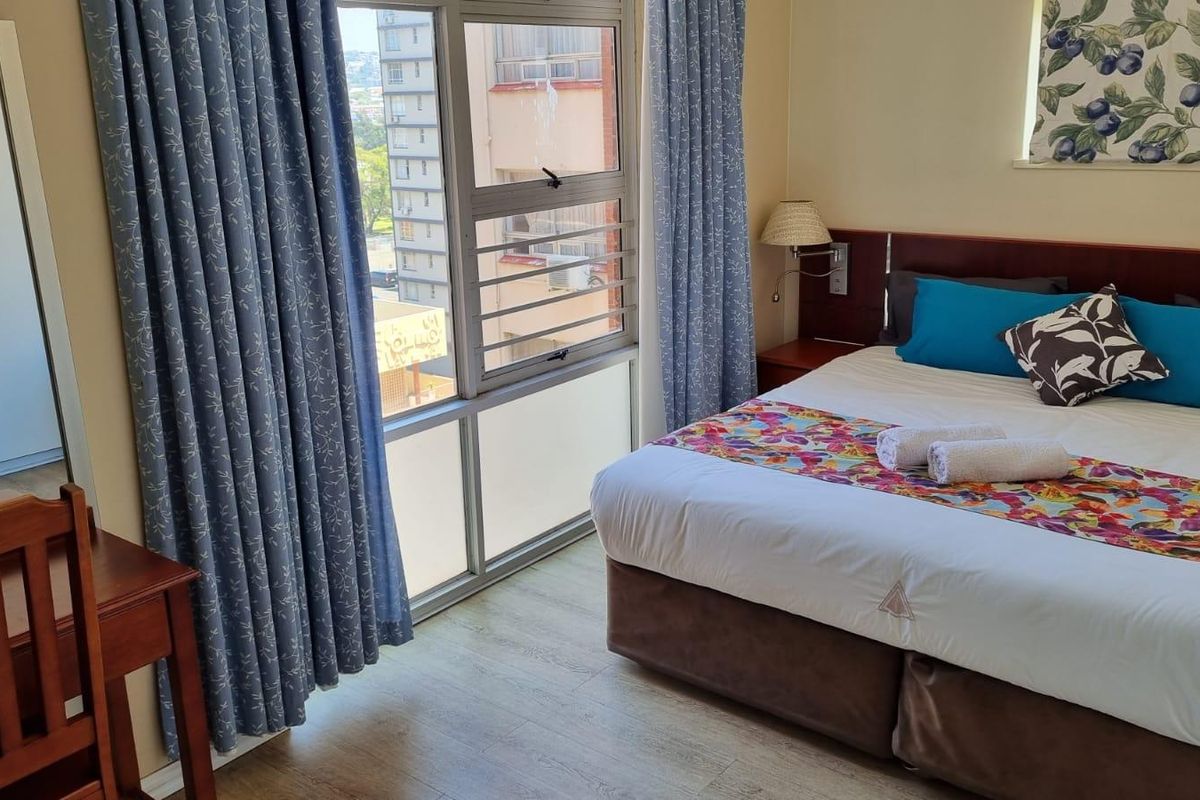 A bedroom featuring light wood-look flooring, a built-in wardrobe, and large windows providing natural light and an urban view.