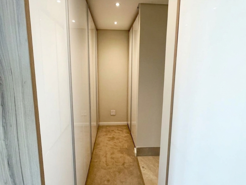 A clean, modern hallway featuring extensive built-in floor-to-ceiling cupboards with high-gloss white finishes and recessed downlighting.