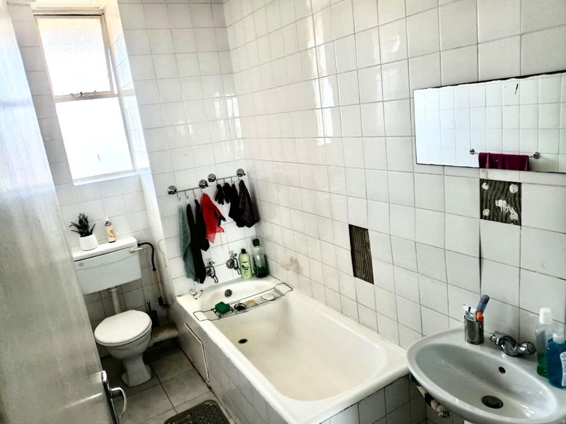 A dated bathroom featuring a bathtub, pedestal basin, and toilet, with white wall tiling and visible areas of missing or damaged tiles.