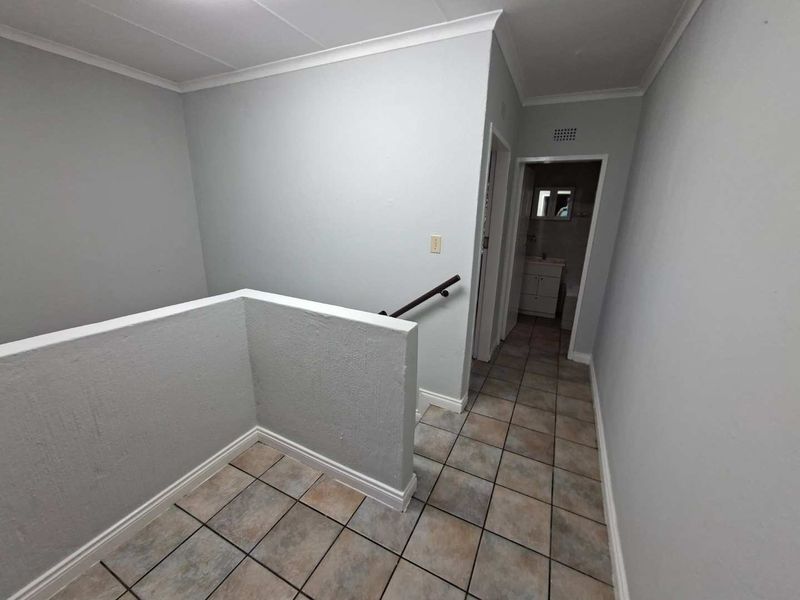 A clean and functional upper-level hallway featuring tiled flooring, a staircase with a handrail, and access to an adjacent bathroom.