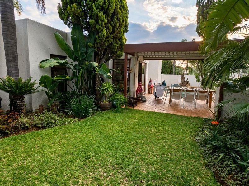 A well-maintained rear garden featuring a lush lawn, mature landscaping, and a covered patio area with brick paving.