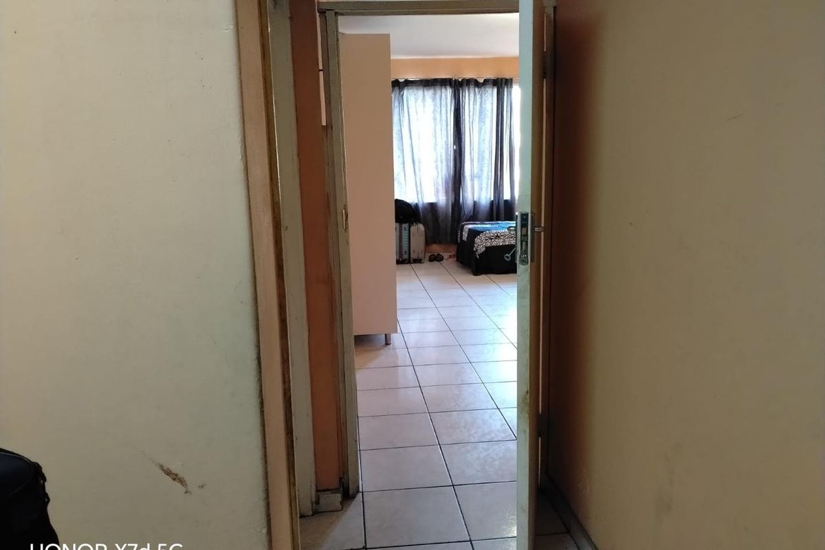 A view through a doorway into a bedroom, featuring tiled flooring and showing signs of wear on the surrounding walls and door frame.