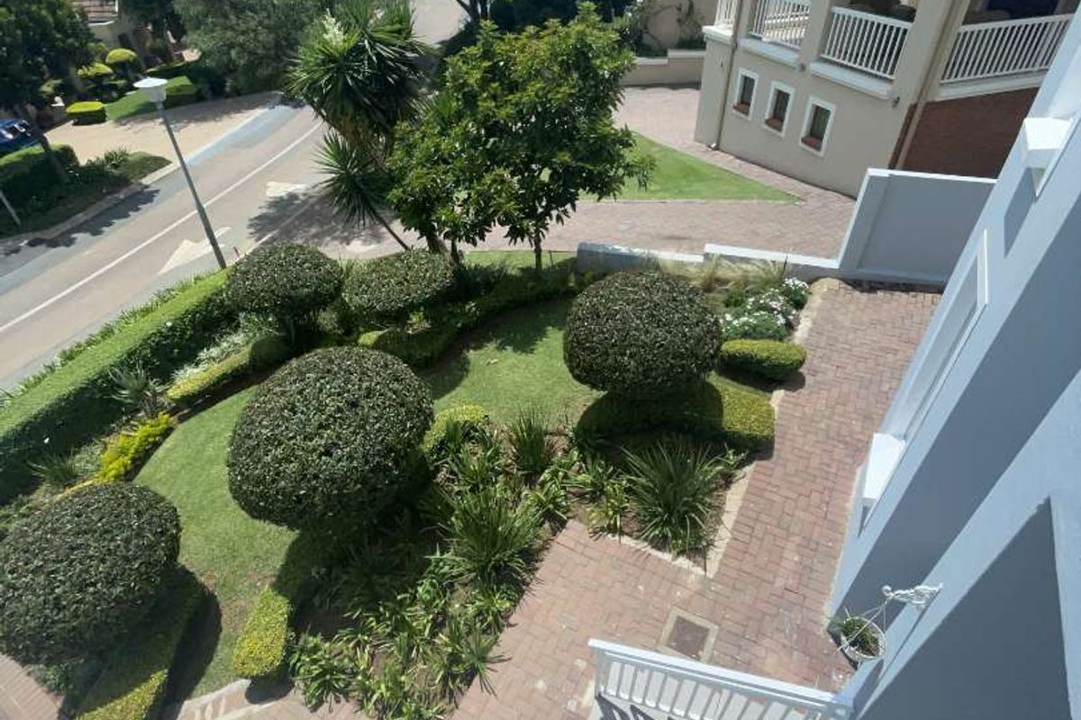 Elevated view overlooking a landscaped garden with manicured shrubs, paved pathways, and surrounding residential architecture.