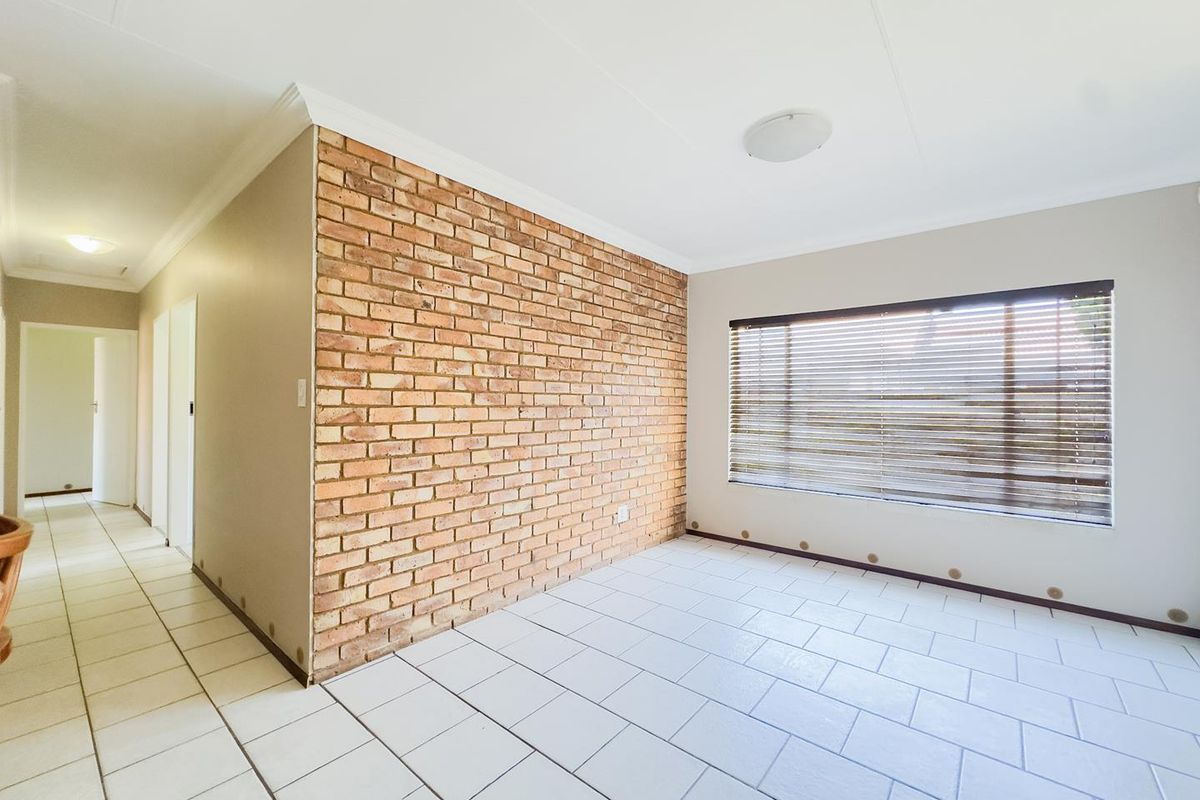 A bright, empty living room featuring a prominent exposed brick feature wall, neutral tiled flooring, and large window with fitted blinds.