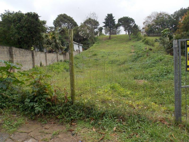 A sloped residential vacant land plot featuring perimeter walling and wire fencing, currently covered in overgrown vegetation.