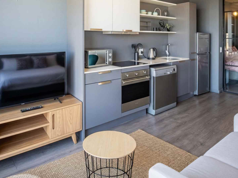 Modern open-plan apartment featuring a compact kitchen with built-in appliances, sleek cabinetry, and a glass-partitioned bedroom area with wood-look laminate flooring.