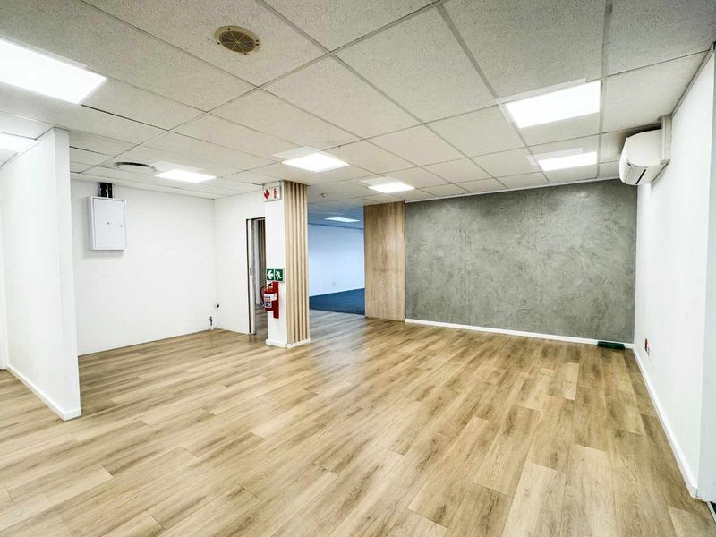 A bright, modern open-plan office space featuring wood-look flooring, a suspended ceiling with LED lighting, and a contemporary textured feature wall.