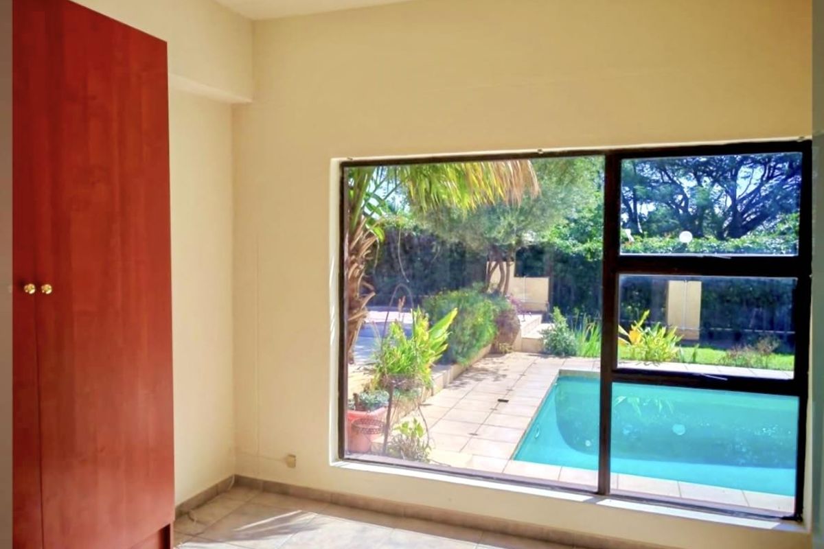A bright, empty bedroom featuring a built-in wardrobe, tiled flooring, and a large window offering a view of the outdoor pool area.