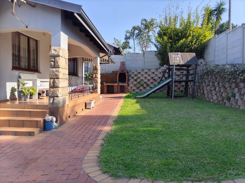 Rear garden area featuring a brick-paved patio, stone-clad pillars, and a built-in brick braai, complemented by a lawn and retaining wall.