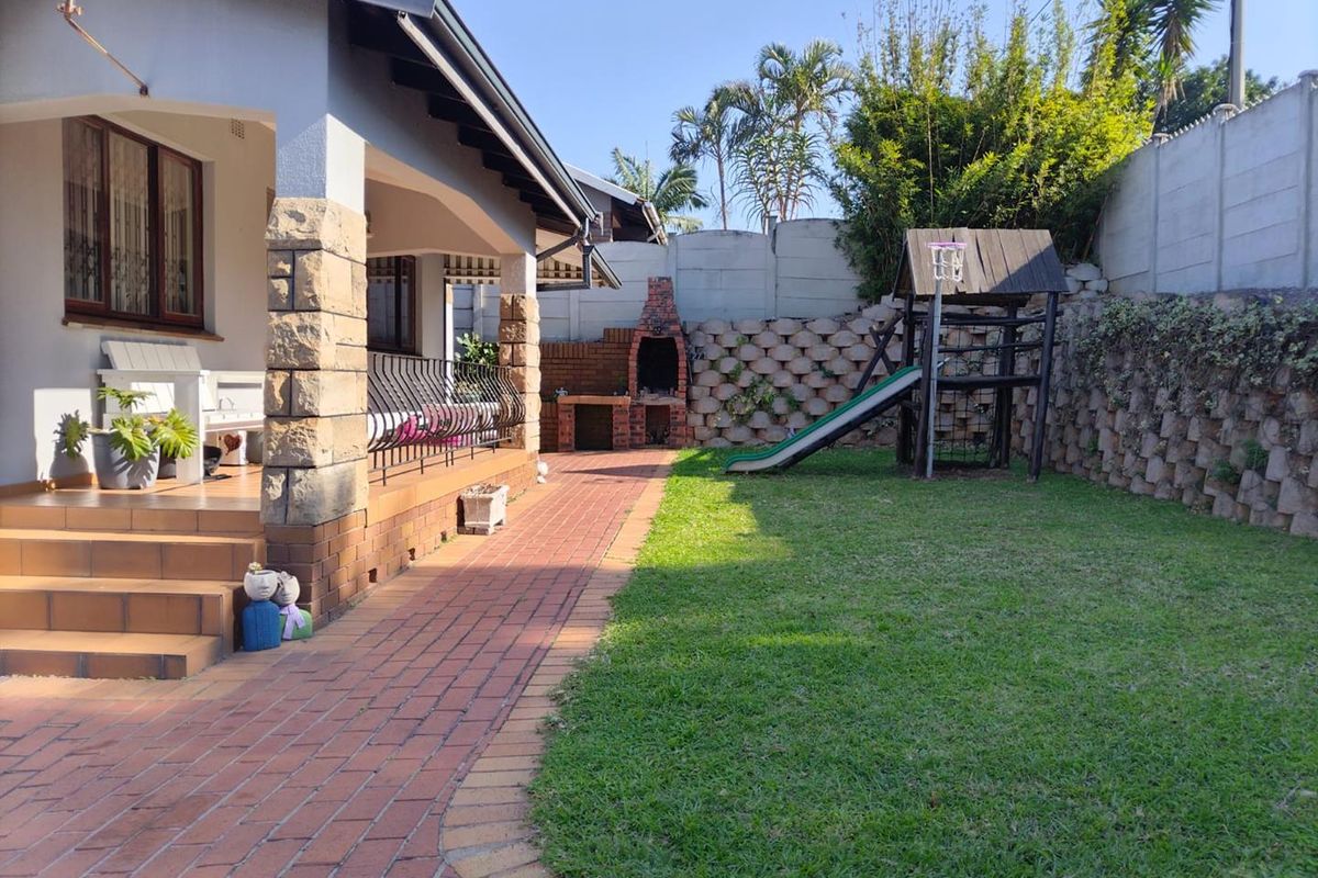 Rear garden area featuring a brick-paved patio, stone-clad pillars, and a built-in brick braai, complemented by a lawn and retaining wall.