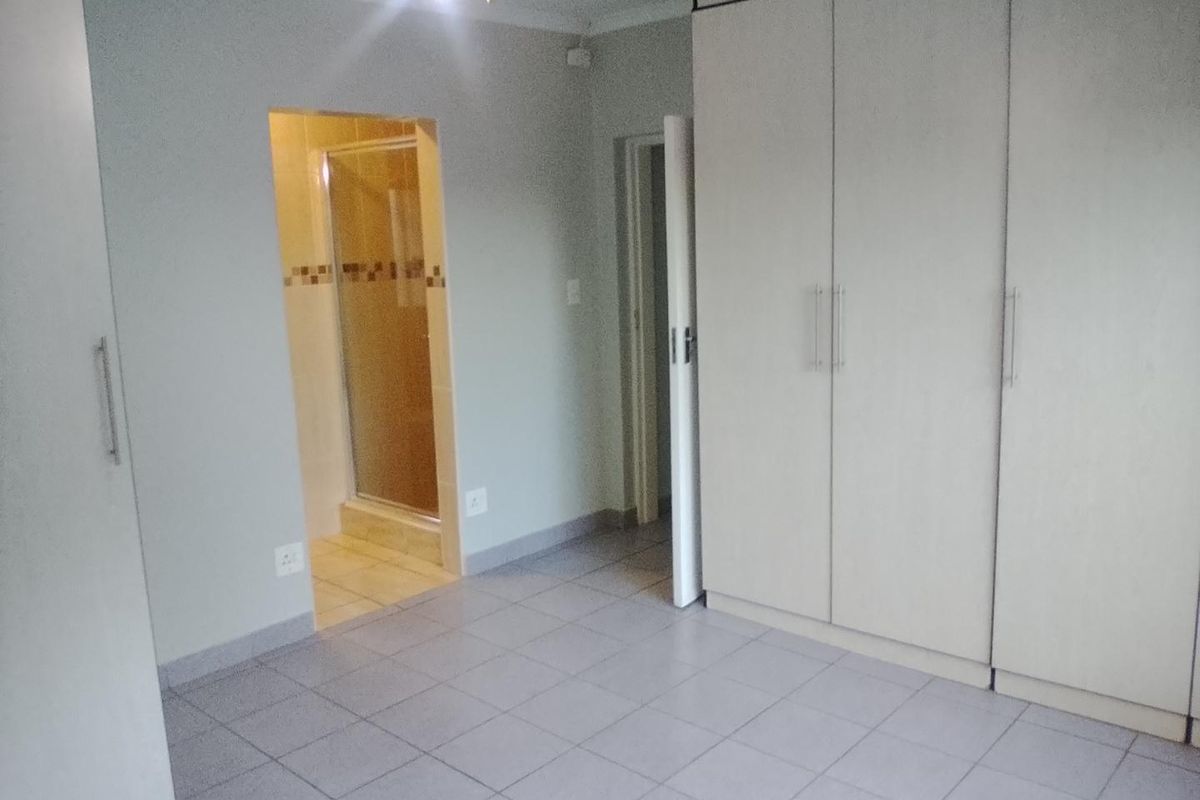 A spacious bedroom featuring neutral tiled flooring, extensive built-in cupboards, and direct access to an en-suite bathroom with a shower.