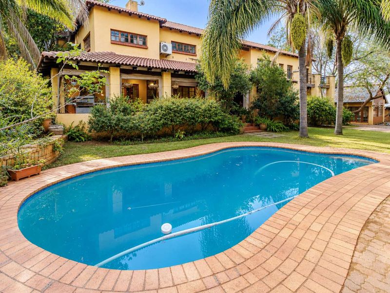A well-maintained rear garden featuring a swimming pool with brick coping, set against a two-story home with a covered patio and terracotta roof tiles.