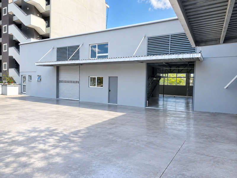 A modern industrial-style exterior space featuring a large concrete paved area, roller shutter door, and a covered entrance with a steel staircase.