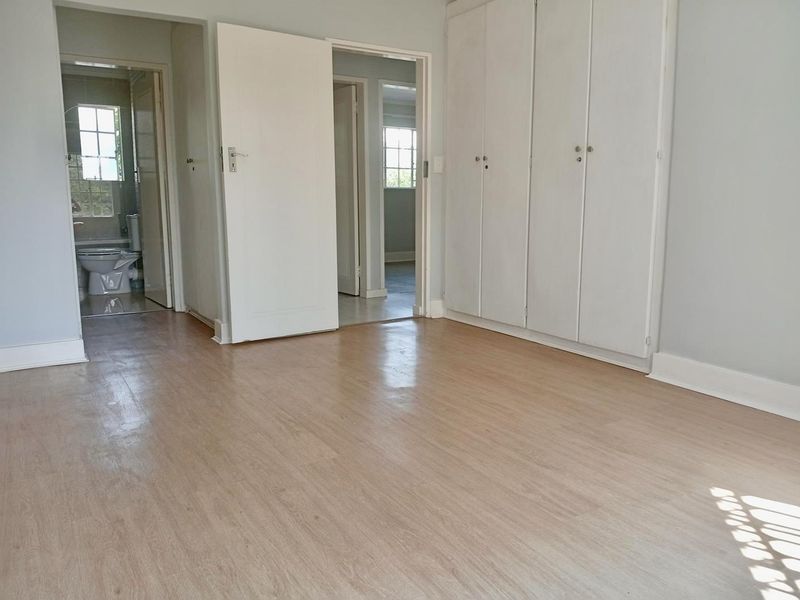 A bright, empty bedroom featuring light-toned laminate flooring, built-in cupboards, and direct access to an adjacent bathroom and hallway.