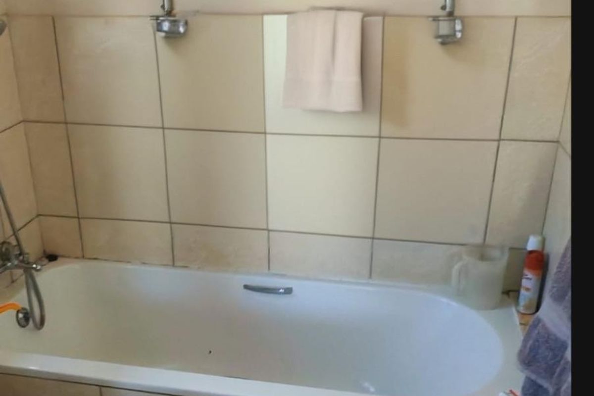 A functional bathroom featuring a built-in bathtub with tiled splashback and chrome fixtures, requiring some maintenance to the grout and sealant.