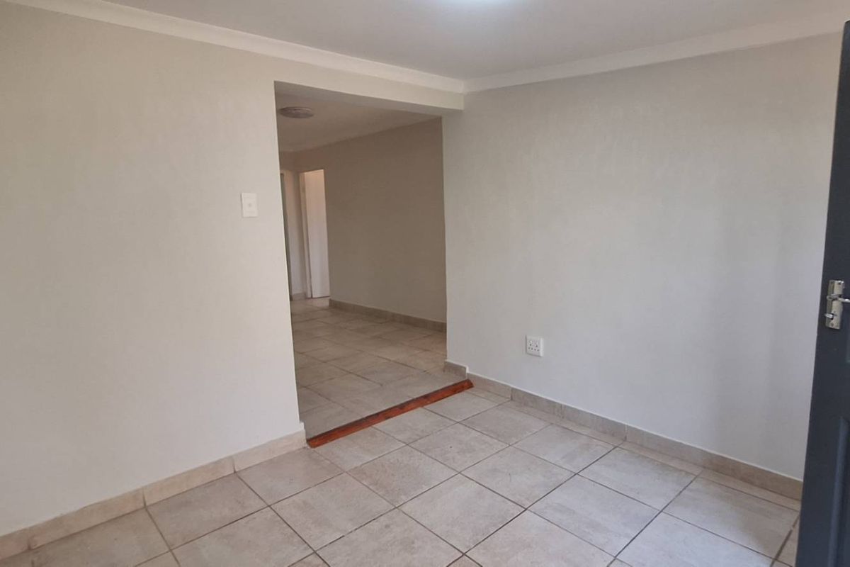 An empty, neutrally painted living area featuring tiled flooring and a coved ceiling, leading into an adjacent hallway space.