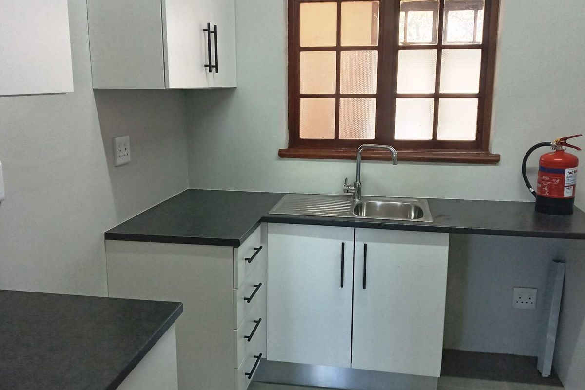 A clean and functional office kitchenette featuring white cabinetry, dark laminate countertops, a stainless steel sink, and a timber-framed window.