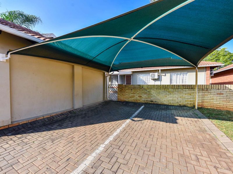 A paved outdoor parking area featuring a protective shade sail structure and a brick boundary wall.