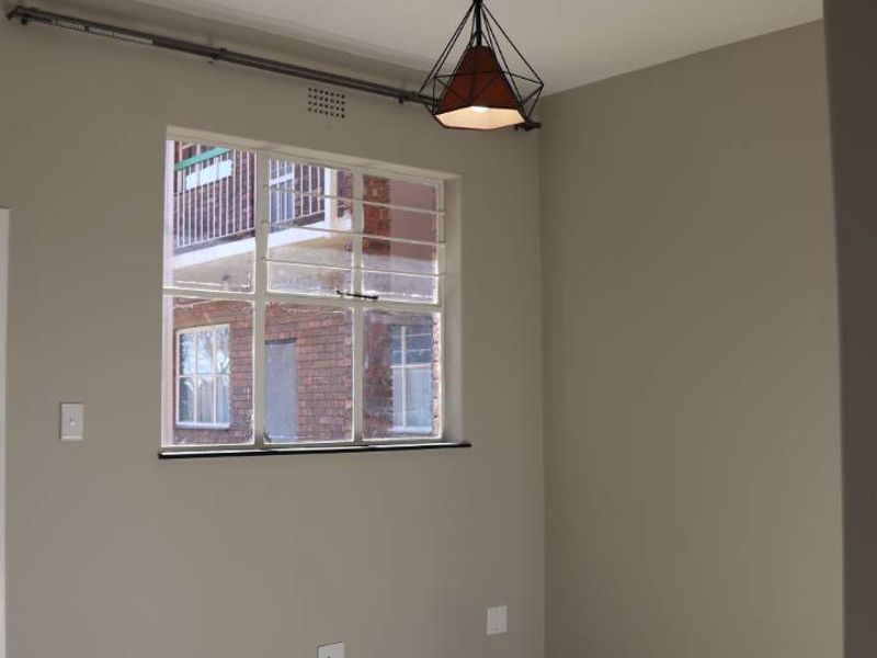 A bright, empty living area featuring neutral wall tones, tiled flooring, and a large steel-framed window providing natural light.