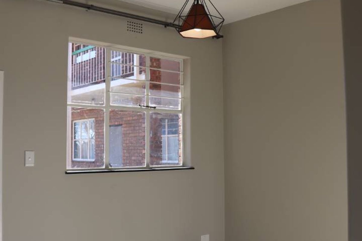 A bright, empty living area featuring neutral wall tones, tiled flooring, and a large steel-framed window providing natural light.