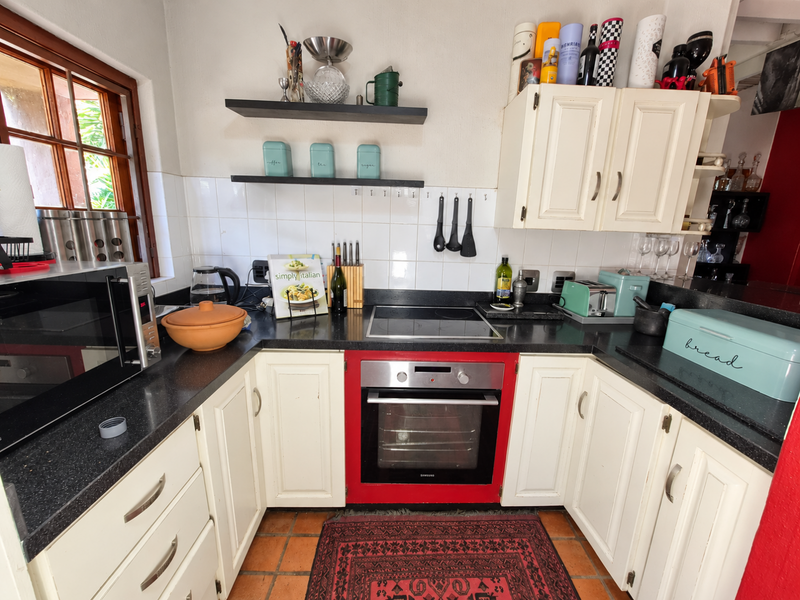 Compact kitchen featuring white cabinetry, black stone countertops, built-in oven with red trim, electric hob, and traditional terracotta floor tiles.