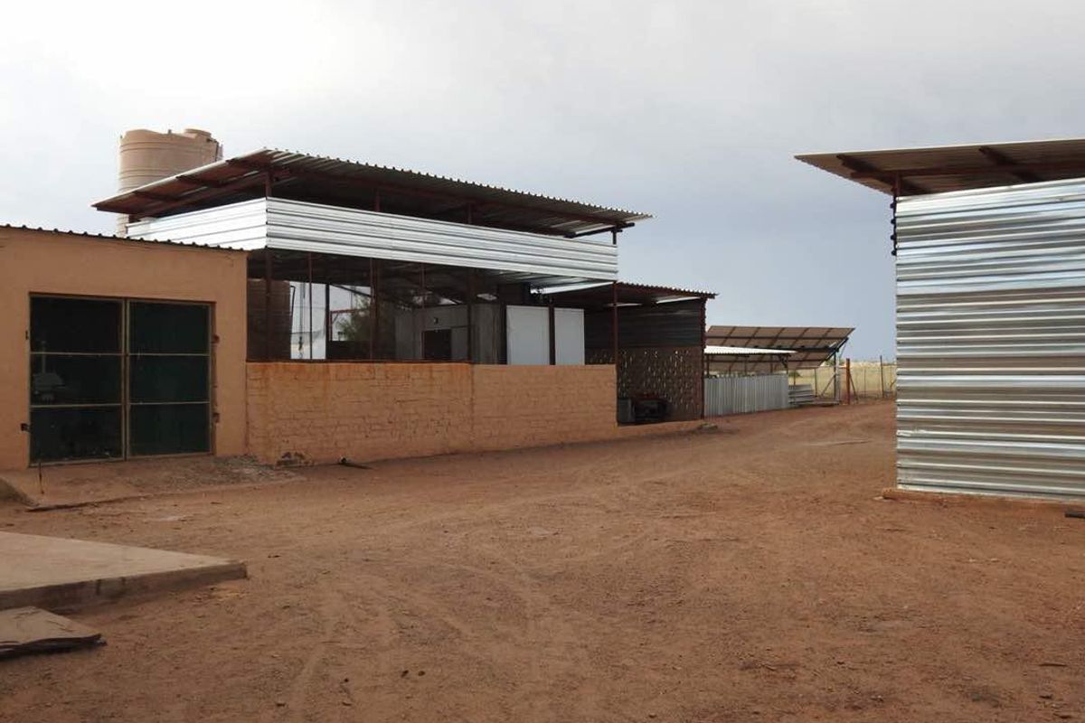 Exterior view of farm utility buildings featuring corrugated metal roofing, brick masonry walls, and a water storage tank on a dirt yard.