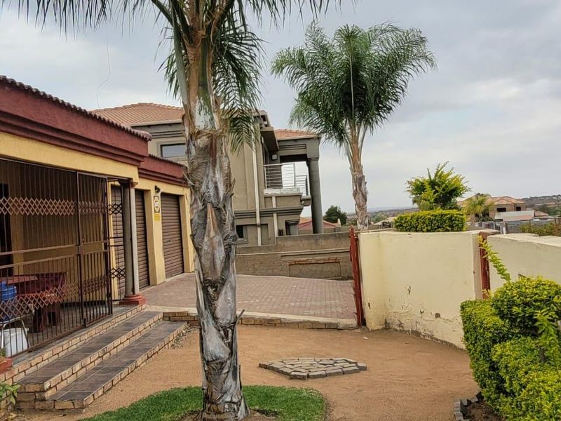 Front exterior view featuring a paved driveway, security gate, tiled patio area, and boundary walling with established palm trees.