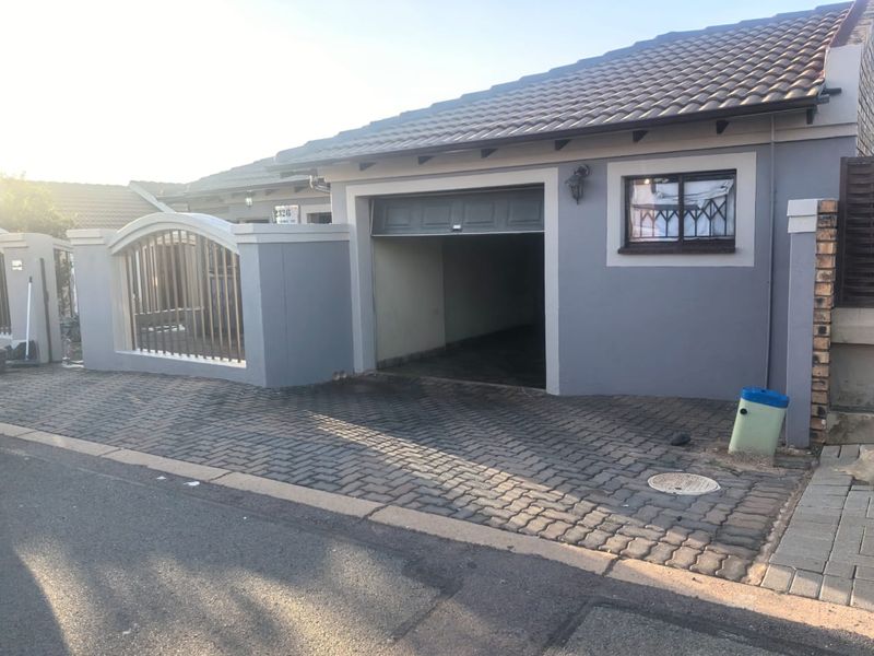 A street-facing view of a residential property featuring a paved driveway, a secure boundary wall with metal gate, and a single garage entrance.