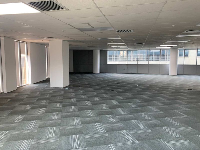 A spacious, open-plan office area featuring carpet tiles, suspended ceilings with recessed lighting, and large windows providing ample natural light.
