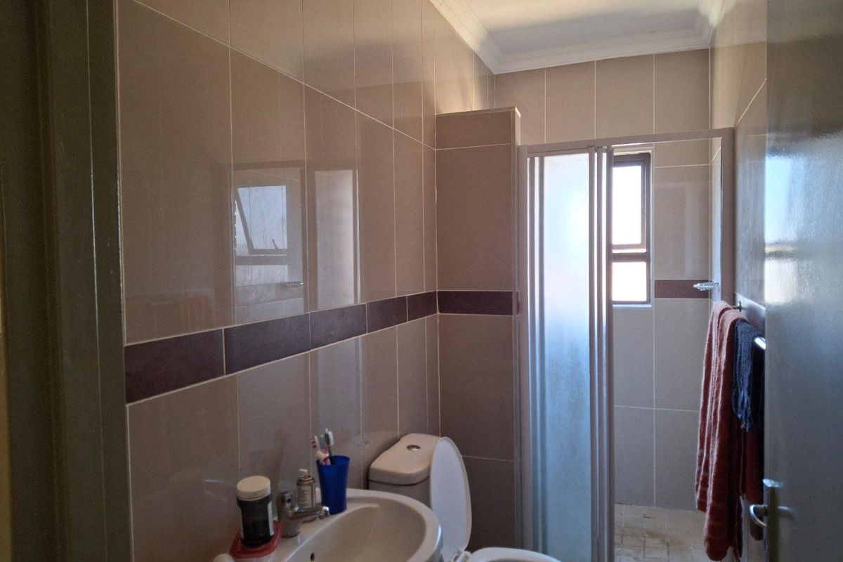 A functional bathroom featuring neutral wall tiling, a pedestal basin, a toilet, and a glass-enclosed shower stall with a small window.