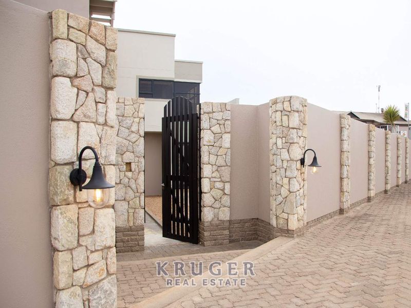 A modern front entrance featuring a secure pedestrian gate, stone-clad pillars, and exterior wall lighting, set against a neatly paved driveway.